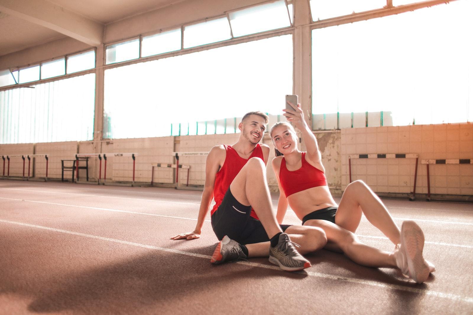 Cheerful young sportsmen smiling and taking selfie on smartphone while sitting on floor and resting after training