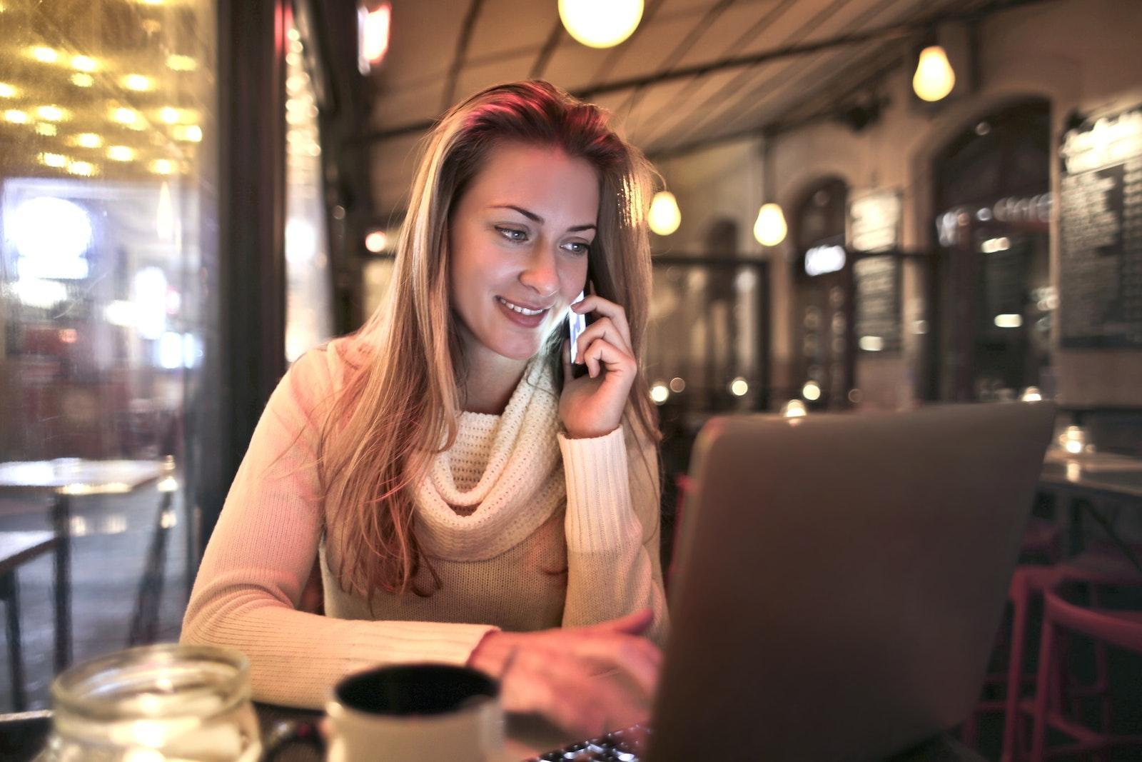 Content female wearing warm sweater sitting at table in cozy cafe and speaking on cellphone while resting in evening and browsing netbook