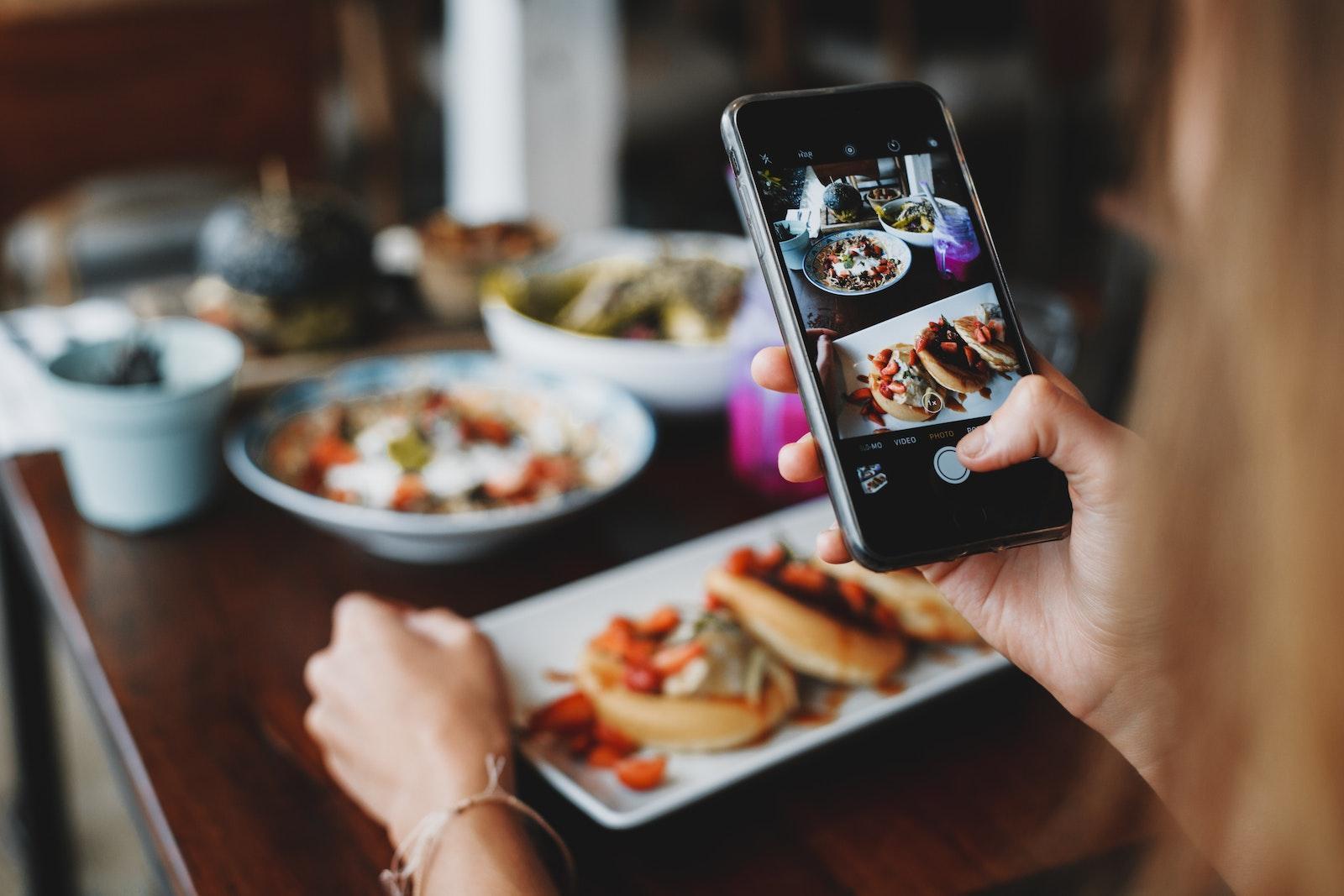 Crop anonymous female taking photo of various tasty dishes on smartphone sitting at table in modern cafe