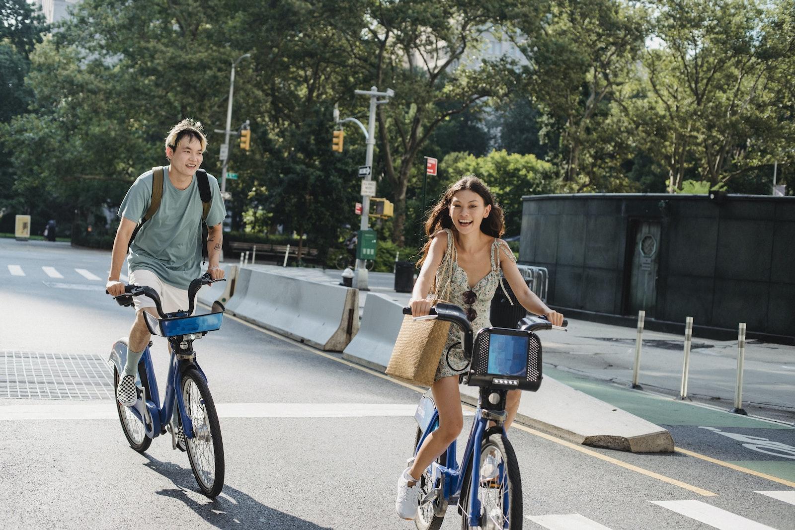 Cheerful diverse couple laughing and riding bicycles on asphalt road in city in sunny day