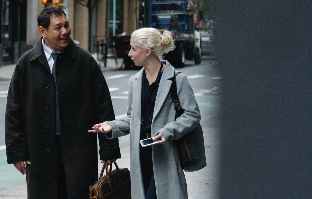 Young woman with blond hair in stylish coat speaking with positive middle aged ethnic male on street
