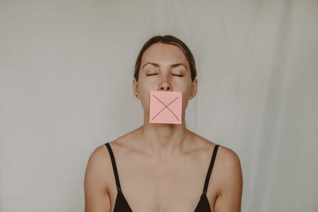 Young slender woman with closed eyes and mouth covered with sticky note showing cross on white background