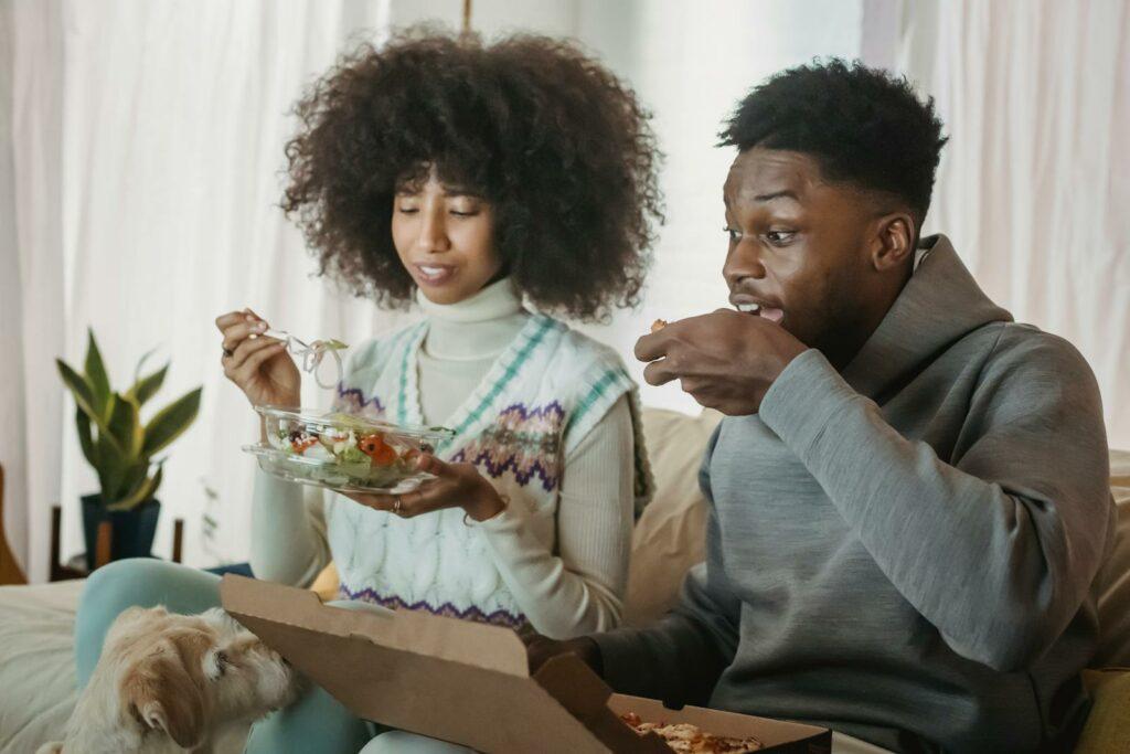 Positive young hungry African American couple with dark curly hair in casual clothes eating appetizing delivered pizza and salad while resting together on sofa near curious dog at home