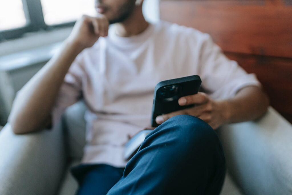 Crop unrecognizable male in casual clothes browsing cellphone while sitting in comfortable armchair in light room near window at home