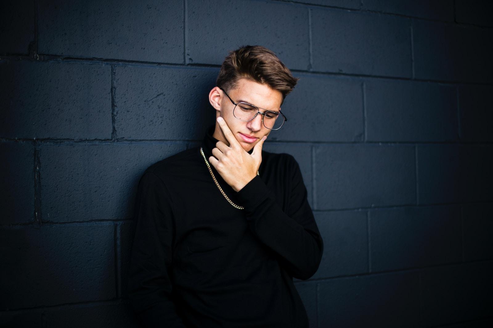 man leaning on wall while looking down wearing eyeglasses and necklace with right hand on chin